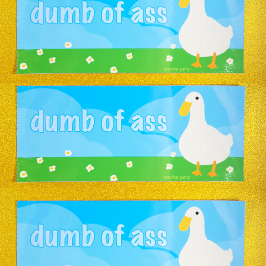 Three bumper stickers sit on a golden sparkly background. The sticker features a cute bird (maybe a goose, or a duck) in a flowery field with a blue sky background. The text on the sticker says, "dumb of ass".