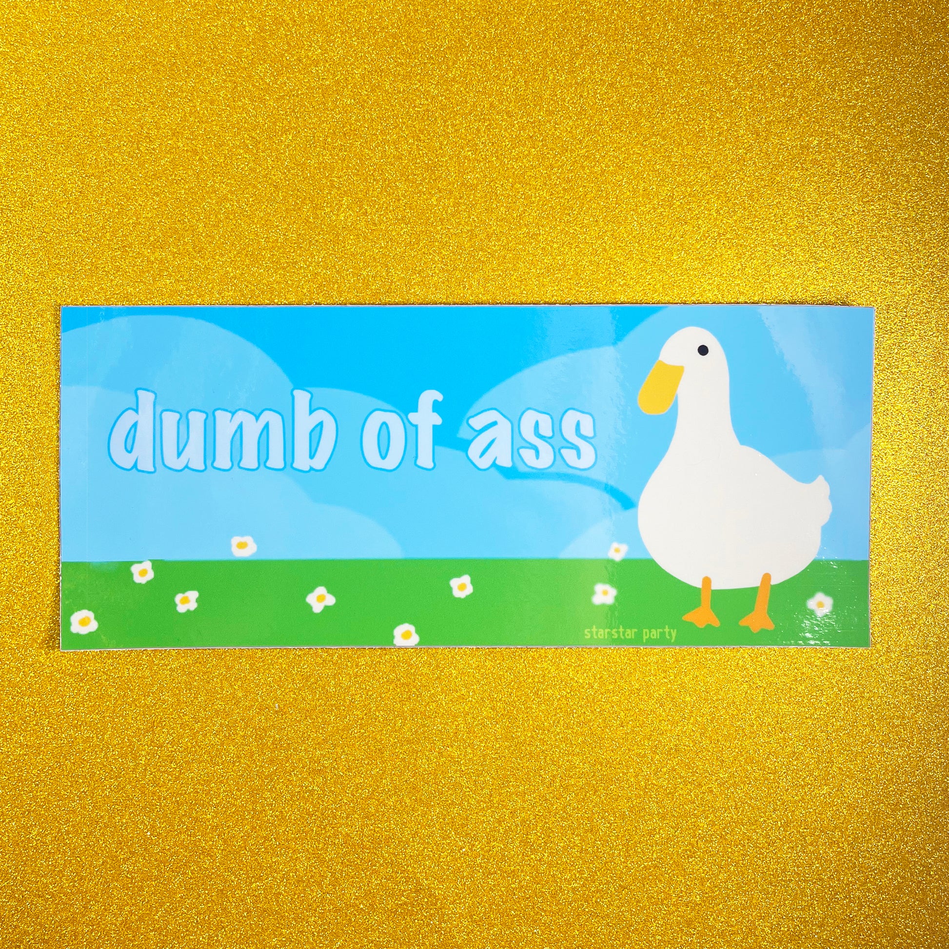 A bumper sticker sits on a golden sparkly background. The sticker features a cute bird (maybe a goose, or a duck) in a flowery field with a blue sky background. The text on the sticker says, "dumb of ass".