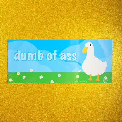 A bumper sticker sits on a golden sparkly background. The sticker features a cute bird (maybe a goose, or a duck) in a flowery field with a blue sky background. The text on the sticker says, "dumb of ass".