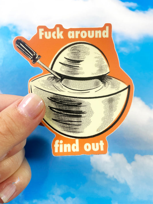 A hand holds up a sticker of an object called the Demon Core. The illustration on the sticker shows the demon core being pried apart with a screwdriver. The font on the sticker says, "Fuck around, Find out".