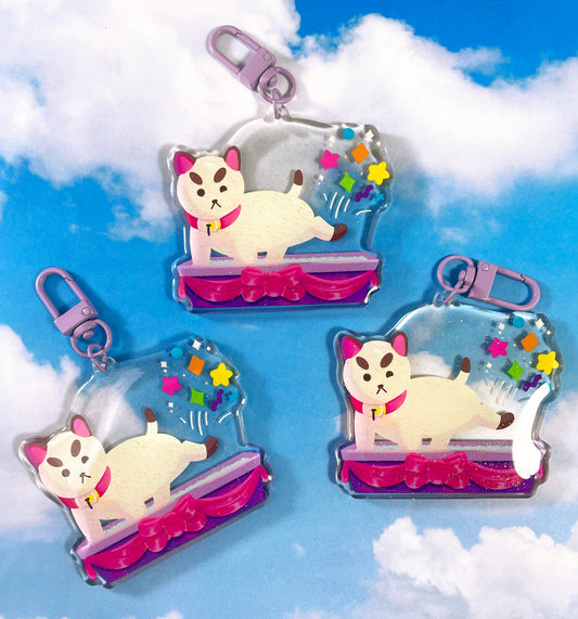 Three keychains sit on a sky print background. The keychains feature a design with a four legged creature that may be a cat or may be a dog. The creature is standing in a purple litter box decorated with a satin pink ribbon. He is kicking colorful shapes into the air behind him. The keychains have a glitter epoxy finish.