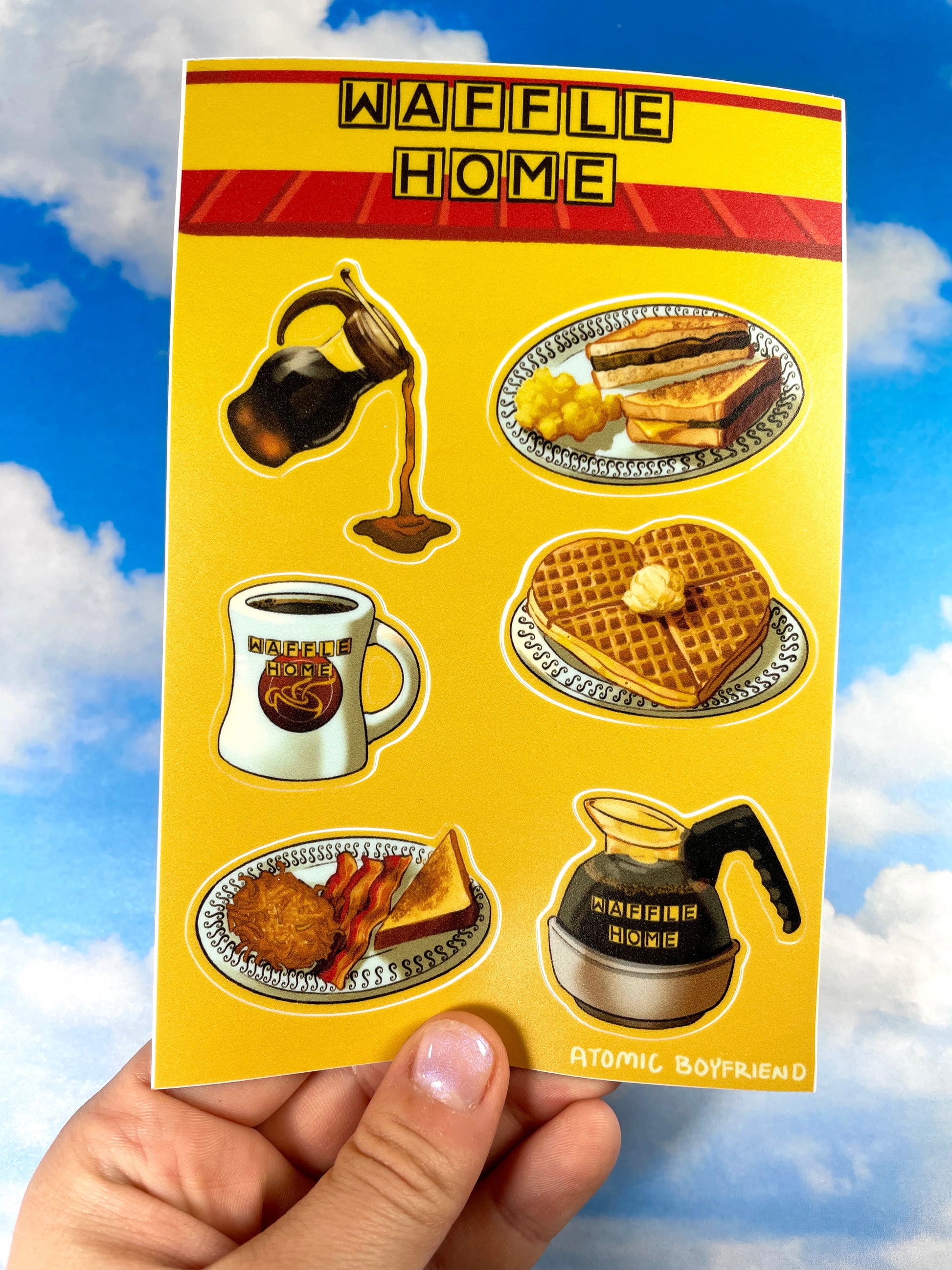 A hand holding a sticker sheet in front of a blue cloudy background. The Sticker sheet features artwork of diner-style breakfast foods. There is a syrup carafe, a heart-shaped waffle, a coffee mug and coffee pot, and two plates of toast, eggs, bacon, and hashbrowns.