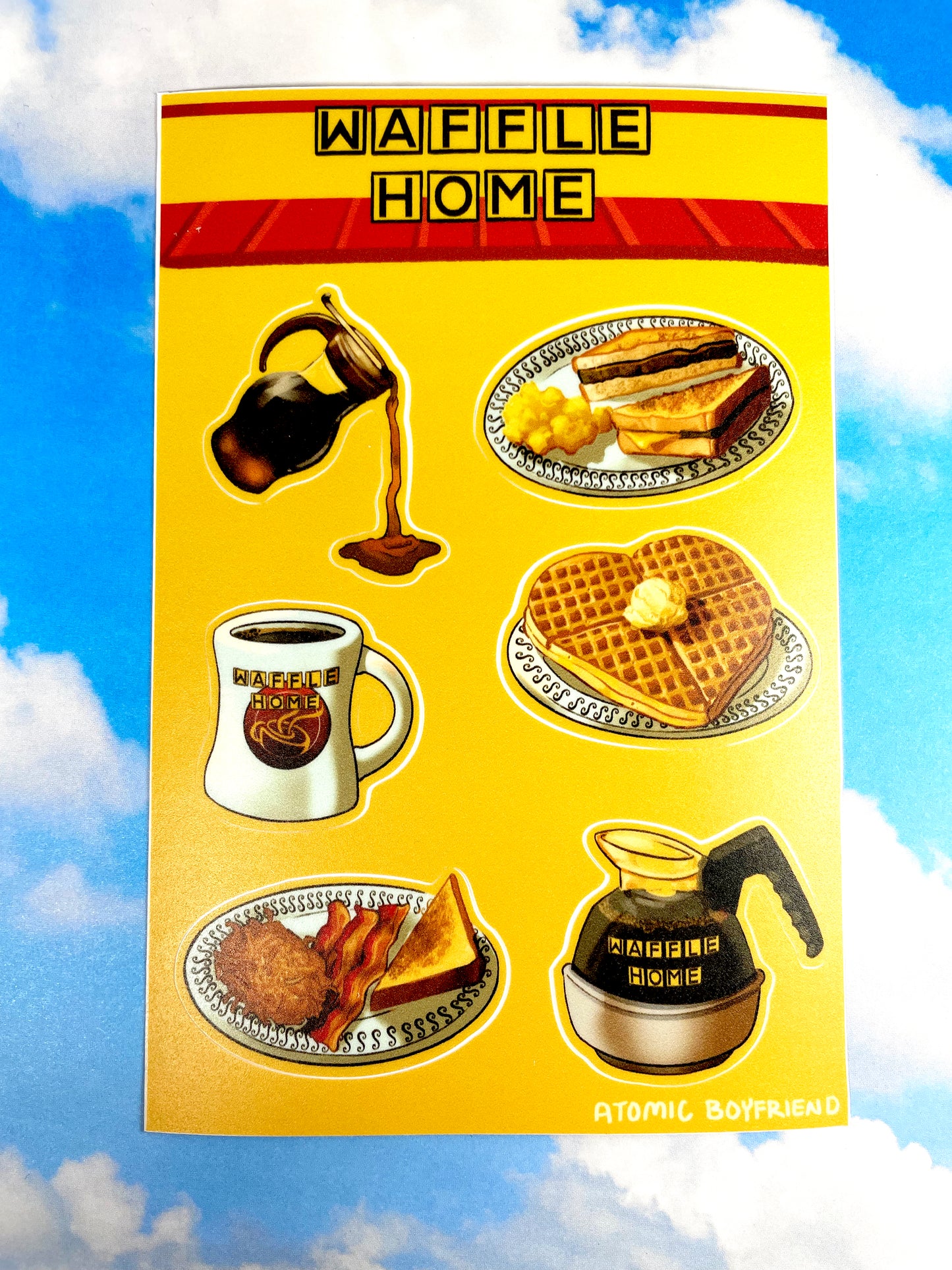 A single sticker sheet on a blue cloudy background. The Sticker sheet features artwork of diner-style breakfast foods. There is a syrup carafe, a heart-shaped waffle, a coffee mug and coffee pot, and two plates of toast, eggs, bacon, and hashbrowns.